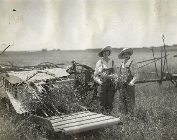 Two women dressed as farmers stand next to a Deering push binder with a cigar and a pipe in their mouths.