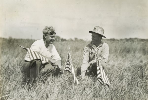 Two men sit in a field along with several small American flags.