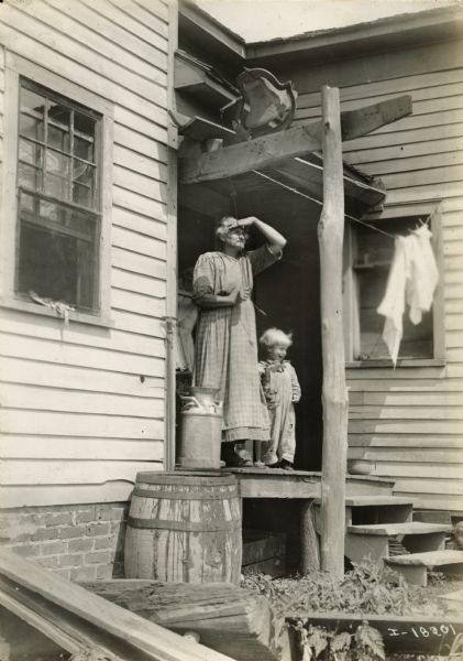 A woman standing on a porch is shielding her eyes from the sun as she is ringing a bell (dinner bell?). A child is standing next to her under a clothesline that is holding a few pieces of laundry.