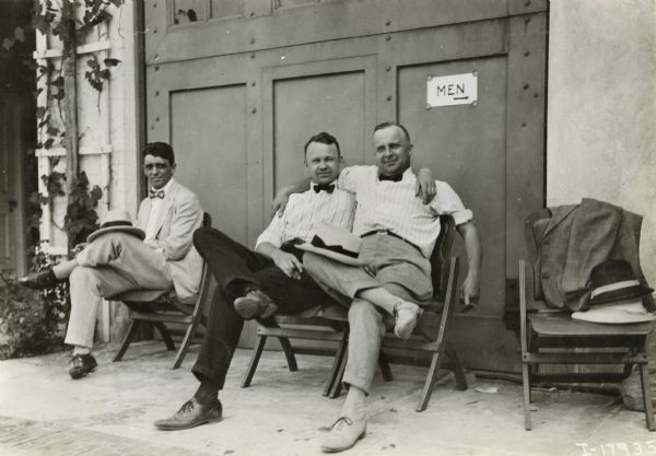 Two men sit side by side on folding chairs with their arms around each other's shoulders. Another man sits in a chair on the left. A sign on the wall behind them reads: "Men" with an arrow pointing off camera.