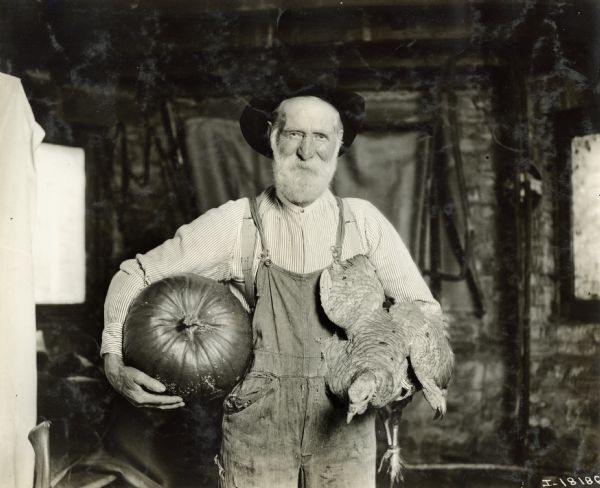 An elderly man, possibly a farmer, holds a turkey and a pumpkin under each arm.