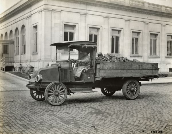 A man is driving an International Model G-61 truck with a bed full of coal for the Clyde Barber Coal Company. Bicycles are leaning against a stone building in the background.