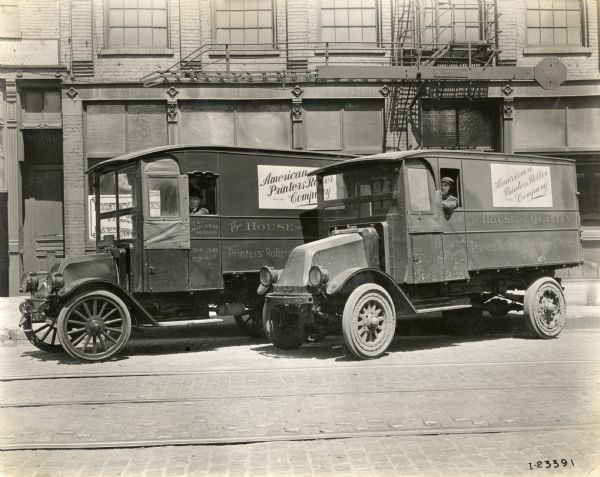 Truck on the left is a Model H or Model F, the truck on the right is an International Model G (Later known as a Model G-61) truck. There is a truck driver in each truck and they are parked on a cobblestone city street. The trucks were operated by the "American Painters' Roller Company."