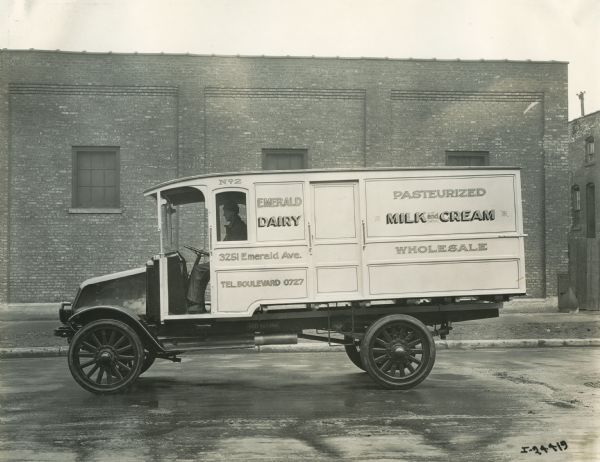 The driver sits behind the wheel of an International Model G-61 truck. The truck was operated by the Emerald Dairy Company, specializing in "Pasturized Milk and Cream." The business was located at 3251 Emerald Avenue.