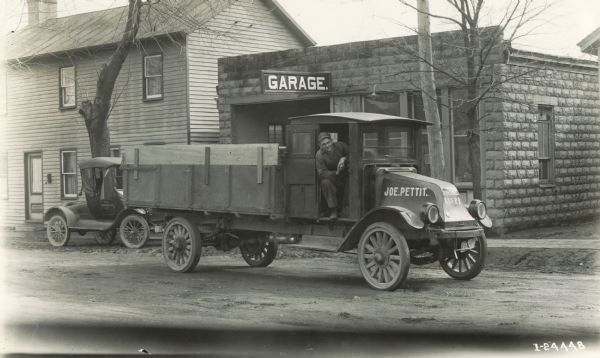 A man leans out of the cab of an International Model G-61 truck parked on an unpaved street in front of a building identified as "garage." The truck has the name "Joe Pettit" painted on it.