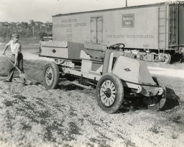 A man, probably John S. Taylor, shoveling gravel into an International Model H Truck. The truck is parked near a railroad box car. Photograph taken by Bergert-Houston.