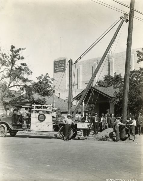 A group of men dressed in suits, ties and hats watch as the San Francisco Electric Department installs a large wooden electric pole. The Electric Company workers wear overalls and more casual shirts while operating an International Model G-61 truck that has been rigged with a crane. A sign for the "Strand Theatre" can be seen in the background.