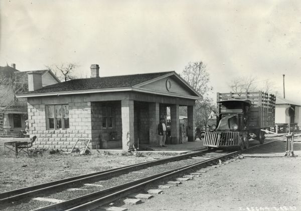 A man stands at a small, run-down station while another man sits in an International Model G or 61 truck that has been rigged with railway gears, wheels and a large wooden bed for the transportation of goods.