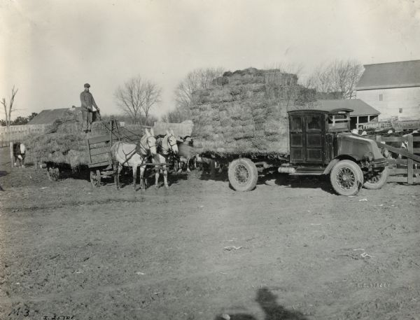 A man stands on a hay-filled bed of a horse-drawn cart on the Peitzman Plain View Stock Farm. Next to the man the bed of an International Model G or 61 truck has been filled with bundles of hay.