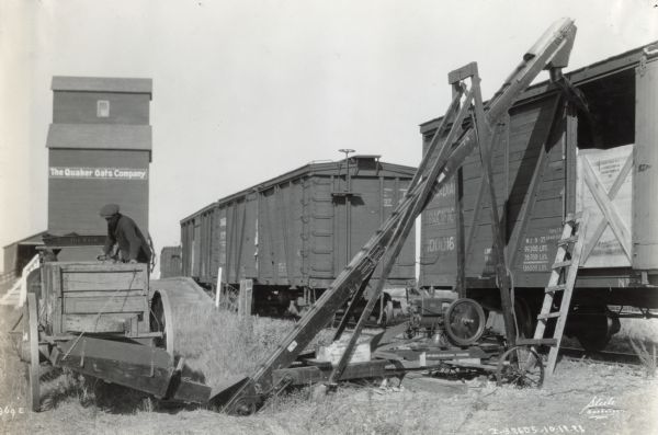 A man uses a McCormick-Deering Kerosene engine to run a conveyor to load grain onto a train boxcar near Saskatoon, Saskatchewan, Canada(?). A building in the background is labeled "Quaker Oats Company."