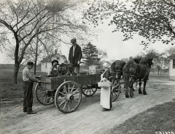 A man stands in a wagon next to an International Harvester engine while other people look on. There is a water tower in the background on the right.
