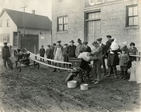 A man surrounded by onlookers is pouring a bag of feed into a grinder powered by an International Harvester stationary engine. A man in the background is wearing an apron advertising "White Satin Flour."