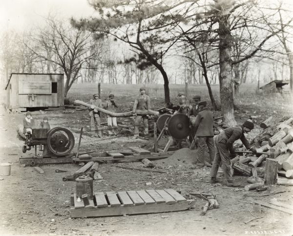 Men use a saw powered by an International Harvester stationary engine. The men are handling logs while two children are looking on.