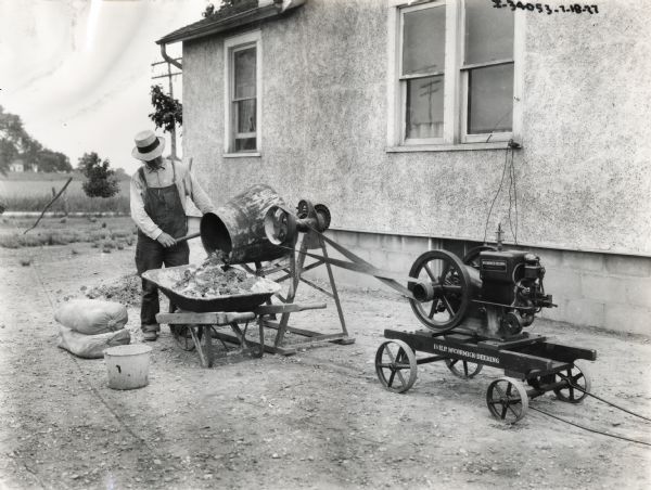 A man pouring mixed concrete into a wheelbarrow. The cement mixer is powered by a McCormick-Deering 1 1/2 h.p. stationary engine.