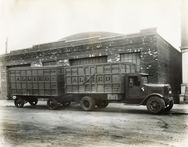 Two men are sitting in an International truck used by the Allied Company. The truck's text reads: "Buchmuller - Eagle Motor; New York, New Jersey, Philadelphia."