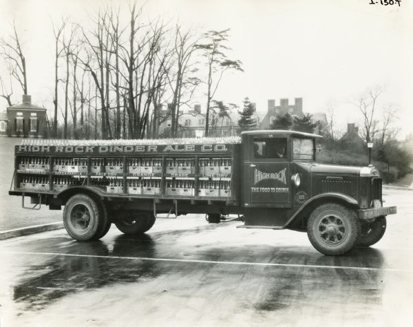 Man driving an International High Rock Beverage truck loaded with bottles of soda. The text on the truck reads: "High Rock-The Food To Drink" and advertises its "Regular Ginger Ale, Sarsaparilla, Pale Dry, Orange, Grape, Lime Dry" flavors.
