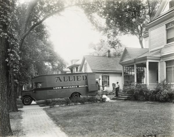 Two men load or unload furniture from an Allied Van Lines moving truck parked in a residential driveway. The truck's lettering reads: "Long Distance Moving; Rock Island Transfer & Storage Co., Agents; Rock Island, ILL."