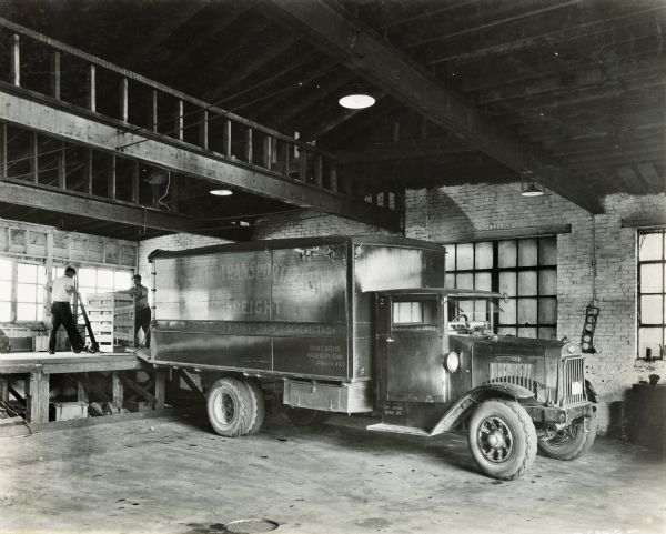 Two men loading a crate onto an International truck used by the Interstate Transportation Company. The truck's lettering reads: "Waterbury-Pittsfield-Albany-Troy-Schenectady; Home Office Waterbury, Conn."