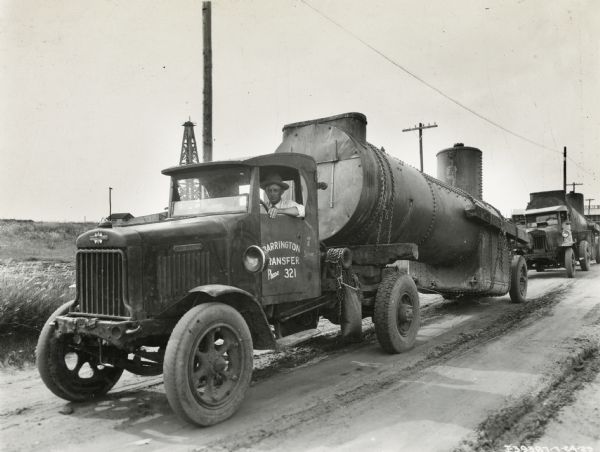 A driver sits in the first of several International heavy-duty oil trucks lined up in front of an oil field. The nearest truck's lettering reads: "Barrington Transfer."
