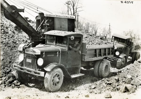 Two men hauling rocks with International truck models W-1. The crane in the background has a sign that reads: "Garrick Bros. Inc. St. Paul, Minnesota".