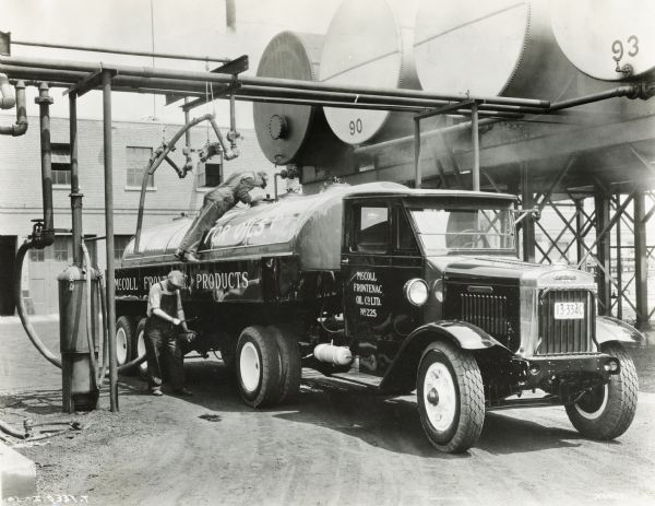 Two men filling an International oil truck used by the Canadian McColl Frontenac Oil Company. Storage tanks and a building are in the background.