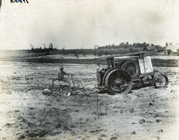 Two farmers use an International tractor to pull a disc harrow through a field. Several farm buildings are in the background.