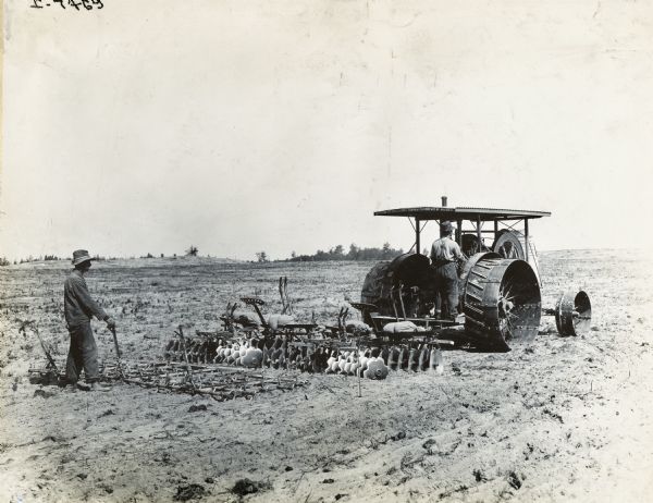 Two men use an International tractor to pull a disc harrow through a farm field.