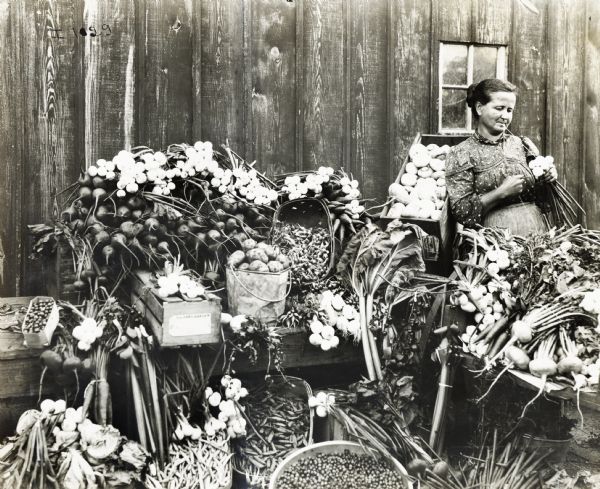 Woman in a printed blouse and skirt tying a bundle of vegetables with teeth and hands. Barrels of vegetables and other produce are set up in a large display.