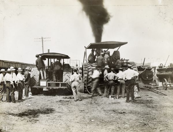 Groups of men in hats, shirts and ties stand around two tractors (one Rumely and one International Harvester) presumably for a demonstration or contest.