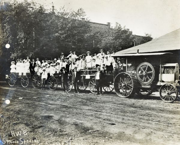 A large group of women, men, young boys and girls in Sunday dress clothes are separated into three wagons pulled by an International tractor. The original caption reads: "H.W.B. Shocco Special Sunday School."