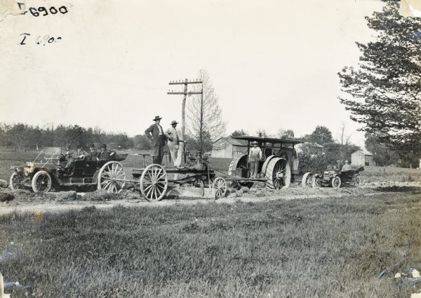 A car with four men in business suits and hats is parked at the side of a road. The men are watching as an International Harvester tractor is pulling a road grader.