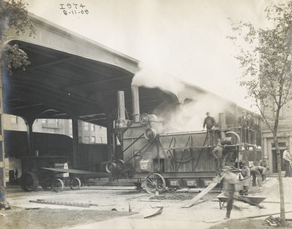 Men running a large machine from the power take-off of an International tractor. The men are working under a metal bridge.