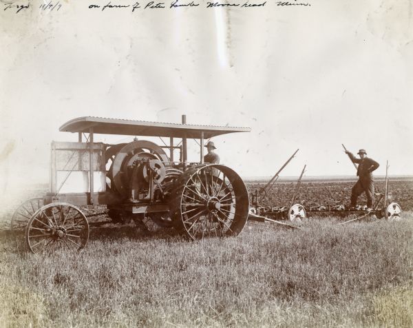 A man operates an International 20 HP friction drive tractor in a field, while another man stands on a trailing plow. The field is on  the farm of Peter Lamb.