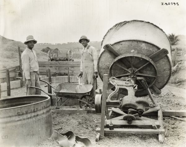 Two men, possibly farmers, work with a wheelbarrow, shovel and cement mixer. One man is dressed in overalls, and both have hats and gloves.