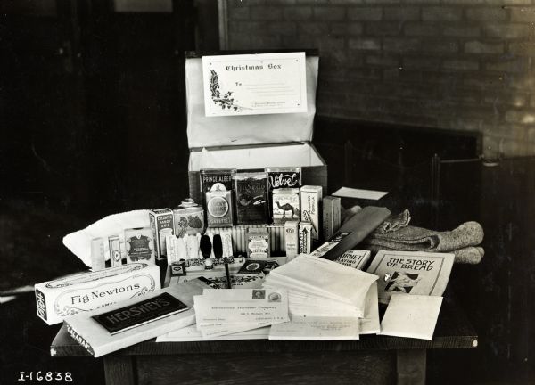 Contents of an International Harvester "Christmas Box", including Fig Newtons, a Hershey's Chocolate Bar, Medicinal Carbolic Soap, Colgate's Handy Grip Shaving Stick, a thimble and thread, a pack of Lucky Strike Cigarettes, Tuxedo Tobacco, a pack of Camel Cigarettes, Wrigley's Spearmint Gum, a pair of wool socks, a pack of playing cards, a book titled "The Story of Bread" and a stack of envelopes. The box was likely intended for U.S. soldiers during World War I.
