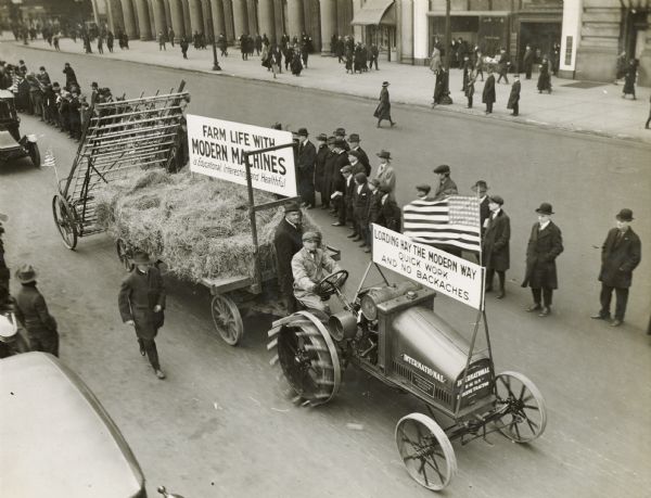 A man pulls a wagon full of hay and a hay loader with an International 8-16 tractor. On both the car and the cart two large signs read: "Loading Hay the Modern Way; Quick Work and No Backaches" and "Farm Life with Modern Machines is Educational, Interesting and Healthful." The display appears to be part of a parade, and people line the street as it goes by.