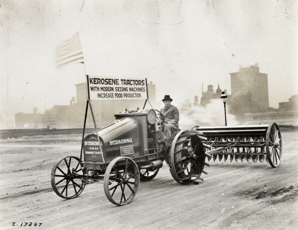 A man drives an International 8-16 HP tractor on a city street with a sign that reads: "Kerosene Tractors with Modern Seeding Machines; Increase Food Production." The tractor is pulling a grain drill.