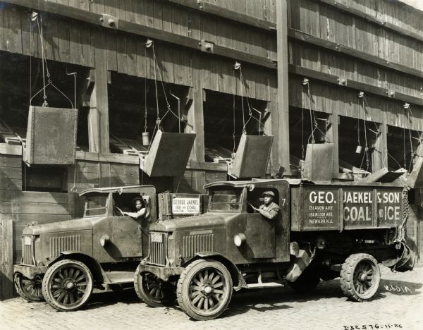 Two drivers sit are sitting their International Model "63" trucks, parked underneath coal chutes. The drivers worked for George Jaekel and Son's Coal and Ice Company in New York.