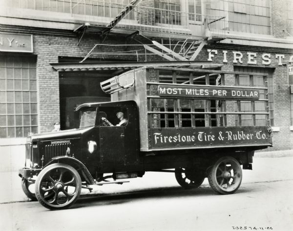 A man is sitting at the wheel of an International Model "63" truck owned by the Firestone Tire and Rubber Company. The slogan: "Most Miles Per Dollar" is painted on truck. A Firestone building is behind the truck.