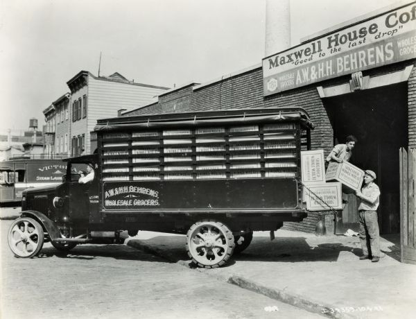 Two men unload (or load) an International Model "63" truck for A.W. & H.H. Behrens Wholesale Grocers. They are handling crates of "Shredded Whole Wheat" at a loading dock.