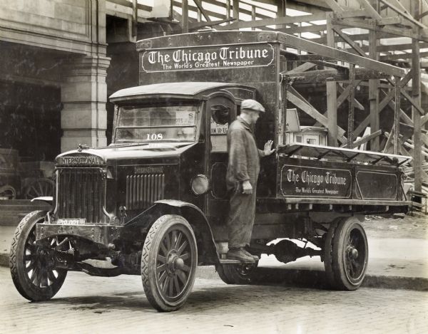 International Model 63 truck operated by the Chicago Tribune Company. The male driver is standing on the truck's running board and looking to the rear. A sign on the truck reads: "The World's Greatest Newspaper." Behind the truck is a large wooden scaffolding structure.