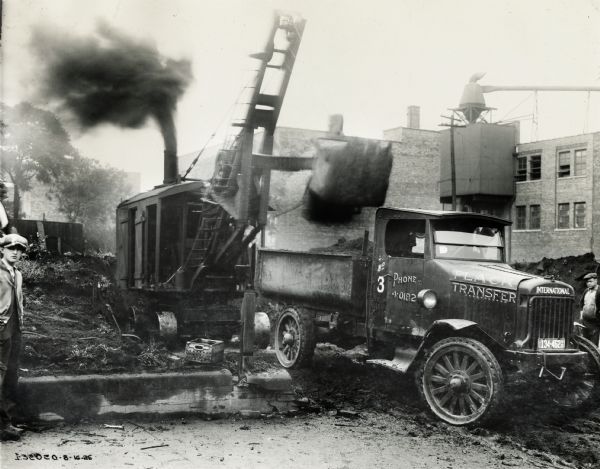 Men loading an International Model 63 truck with a steam shovel, or crane. The truck is operated by the Plack Transfer Company. The truck has been rigged with large front wheels and a metal bed.