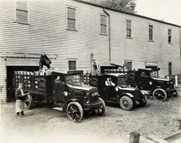 Men loading (or unloading) International Model 63 trucks for the Fountain and Heinzman Company. Slightly elevated view of three drivers sitting in the cabs of the trucks, three employees standing in the filled beds and one man standing to the left of them all holding a clipboard, presumably for inventory.