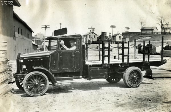 A man sits in the cab of an International Model 63 truck operated by the Woonsocket City Lumber Company. Stacks of lumber are arranged on the ground. Residential homes are in the background.