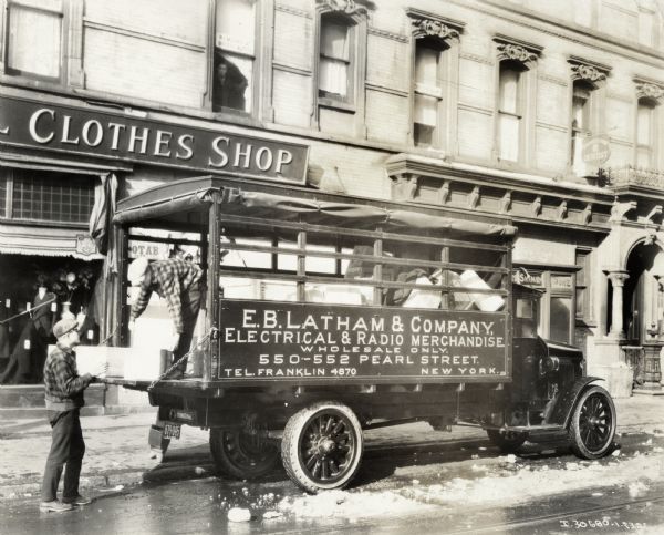 Two men loading merchandise into the covered bed of an International Model 63 truck operated by the E.B. Latham & Company - "Electrical & Radio Merchandise." The truck is parked outside a clothing shop in New York.