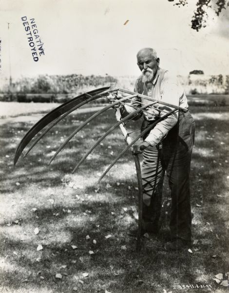 Samuel Hightower(sp?) stands in a grassy yard and holds up a cradle for harvesting grain.