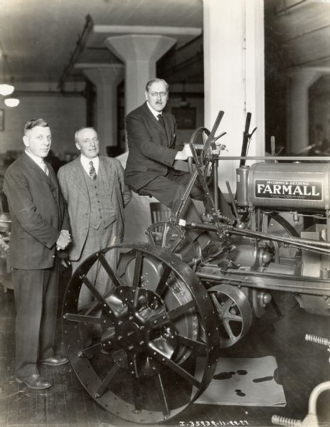 Mr. Brownlie Gout, of Punjab, India, sits on top of a McCormick-Deering Farmall Regular tractor in a showroom, while two men stand to the side.