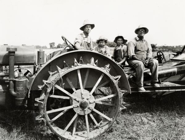 Two men, one of them possibly H.R. Clark, sitting with two boys on a tractor. All of them are wearing overalls and floppy hats.