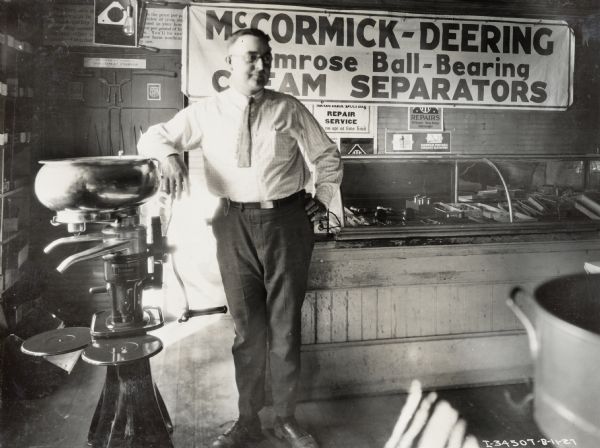 Waldo Hancock leans against a cream separator in his International Harvester dealership, Waldo Hancock and Sons - Hancock Implement Company. On the wall behind Hancock, a large sign reads: "McCormick-Deering Primrose Ball-Bearing Cream Separators".