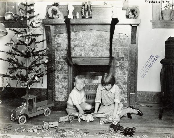 A young boy and girl play with International Harvester toys in front of a fireplace and feather Christmas tree. The toys include a truck, tractor, thresher and wagon. The toys appear to be a series manufactured by Arcade for International Harvester.
